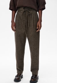 Persoon in hoge bruine wollen broek met een gestrikt trekkoord en donkerbruine leren loafers tegen een rustige witte achtergrond.