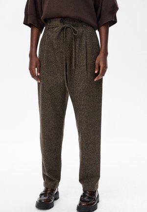 Osoba w wysokich, brązowych wełnianych spodniach z wiązanym paskiem i ciemnobrązowych skórzanych loafersach na tle gładkiego białego tła.