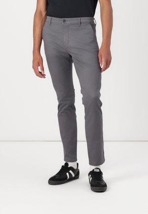 Mann trägt eng anliegende graue Hose, schwarz-weiße Sneaker und weiße Socken vor schlichtem weißem Hintergrund.