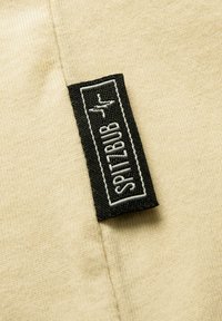 Beiger Baumwollstoff mit einem schwarzen Etikett, auf dem der weiße Text "SPITZBUB" gestickt ist, das prominent im Vordergrund platziert ist.