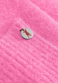 Różowa teksturowana tkanina z okrągłym metalowym znaczkiem przedstawiającym sylwetkę czerwonego psa oraz brązowym sznurkiem do zawieszenia.