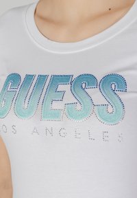 Fehér póló, amelyen a "GUESS" felirat színátmenetes türkiz kivitelben, strasszkövekkel díszítve, az alján pedig kisebb ezüst betűkkel a "LOS ANGELES" felirat található. Sima pamut textúra.