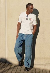 Camisola atlética branca com bandeira italiana, riscas vermelhas e verdes; calças de ganga azuis extragrandes; sapatos pretos; sombra contra uma parede de betão.