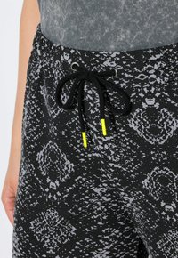 Pantalons de survêtement noirs avec un motif abstrait gris, dotés d'un cordon de serrage noir avec des embouts en néon jaune et d'une taille élastiquée.