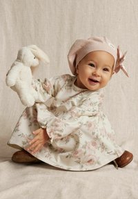 Ljus blommig klänning med volangärmar, beige botten och rosa rosor. Barnet bär en rosa stickad hatt och håller en vit gossedjurkanin.