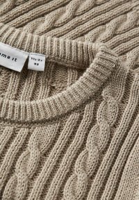 Suéter de punto beige con un diseño de trenzas texturizado. Presenta un cuello acanalado y una etiqueta que indica la talla 1½-2 años.