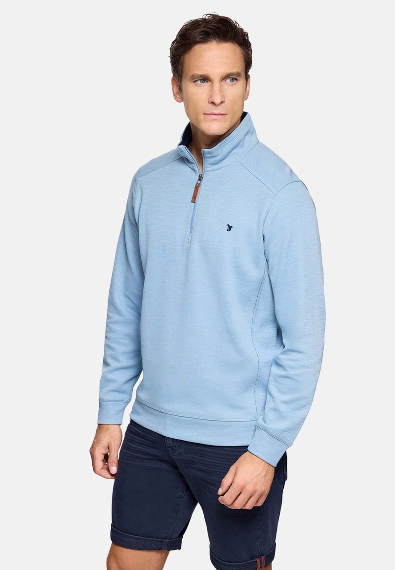 Hombre con jersey de cremallera hasta la mitad color azul claro y pantalones cortos azul marino oscuro, de pie frente a un fondo blanco liso, mirando hacia la derecha.