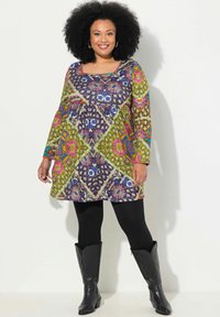 Robe colorée à motifs avec des manches longues, présentant un mélange de verts, de violets et de roses, portée avec des leggings noirs et des bottes.