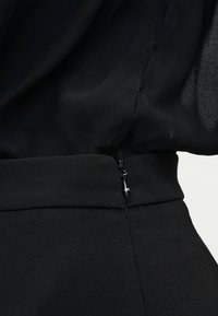 Κοντινή λήψη μαύρου υφάσματος με λεπτομέρεια πλαϊνής φερμουάρ σε ρούχο με την μέση ψηλά, που αναδεικνύει την υφή και το ράψιμο.