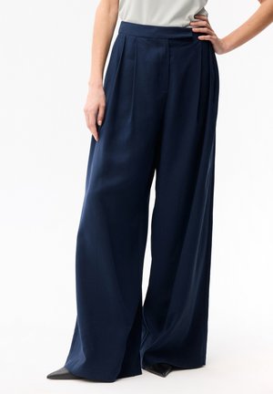 Femme portant un pantalon taille haute à jambes larges bleu marine et des chaussures noires à bout pointu, une main posée sur la hanche et l'autre le long du corps.