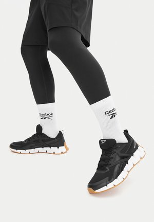 Melni sporta apavi ar fakturētu virsmu, baltas zoles akcentiem un gumijas ārzoli. Valkāti kopā ar melnām legingām un baltiem zeķēm ar logo.