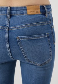 Jeans de mezclilla en azul medio, con un parche de cuero etiquetado como "pantalones perfectos" en la cintura y un solo bolsillo trasero con detalles de costura.