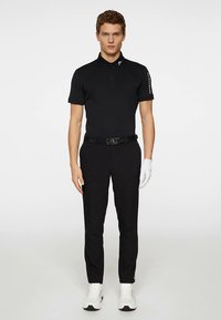 Μαύρο πόλο πουκάμισο με γιακά, κοντά μανίκια και λεπτομέρεια λογότυπου στον ώμο. Συνδυάζεται με μαύρο παντελόνι και λευκά αθλητικά παπούτσια. Χέρι με γάντι.