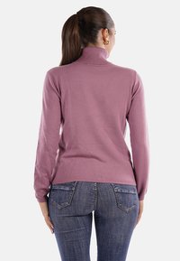 Maglione rosa lavorato a maglia con colletto alto, maniche lunghe e silhouette aderente, abbinato a jeans di denim blu scuro. Texture liscia visibile.