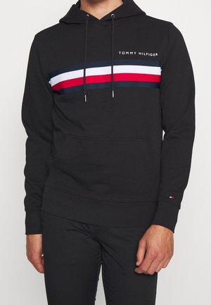 Mężczyzna w czarnej bluzie z kapturem Tommy Hilfiger z granatowym, białym i czerwonym poziomym paskiem na wysokości klatki piersiowej oraz kieszenią kangurką.