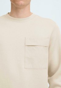 Beige langemouwen shirt gemaakt van zacht stof met een ronde halslijn en een enkele patch zak op de linker borst. Eenvoudige, gladde textuur.