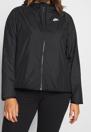 Vrouw die een zwarte Nike zip-up capuchontrui met lange mouwen en zwarte leggings draagt tegen een effen achtergrond.