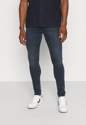 Homme portant un jean skinny foncé, une chemise noire boutonnée et des baskets blanches avec des accents noirs, debout devant un fond uni.