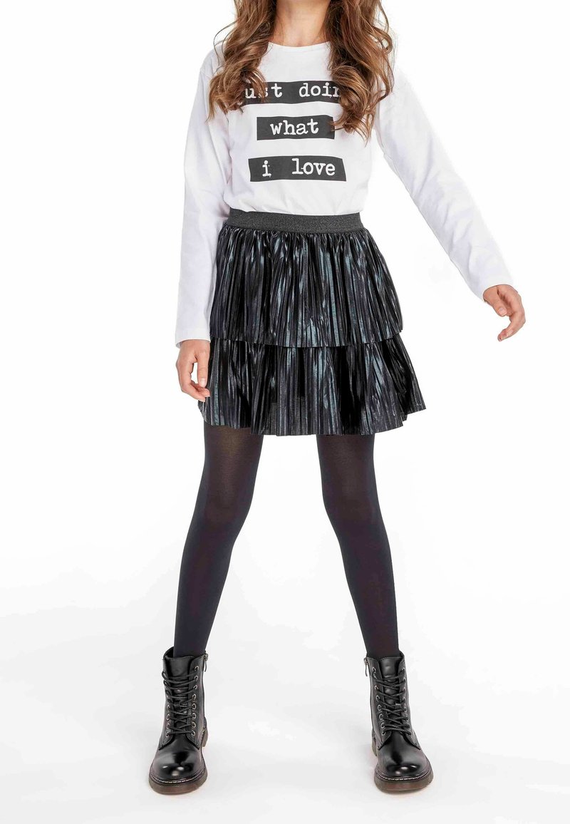 Chemise blanche à manches longues avec texte noir, jupe plissée noire superposée, collants noirs et bottes noires à lacets avec une finition brillante.