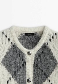 Gros plan d’un cardigan blanc et gris tricoté avec de grands motifs en losange et deux boutons métalliques à l’avant.