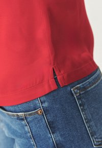 Rode blouse met zijsplitten, gemaakt van gladde stof, gecombineerd met vervaagde blauwe denim jeans met een koperen knoop en contrasterende stiksels.