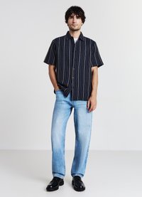 Camicia azzurra a maniche corte con strisce verticali bianche, abbottonata sul davanti, abbinata a jeans straight-leg blu chiaro e scarpe nere. Outfit casual.