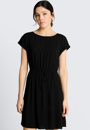 Jeune femme aux cheveux foncés porte une robe noire à manches courtes avec une taille nouée, debout devant un fond gris clair uni.