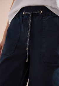 Primer plano de pantalones azul marino con cintura elástica, cordón negro y blanco, y grandes bolsillos delanteros, usados con una camisa blanca por dentro.