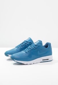 Un paio di sneaker Nike Air Max blu con suole bianche, caratterizzate da tomaia in pelle perforata e ammortizzazione ad aria visibile su una superficie bianca.