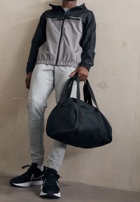 Atletisk outfit med en svart och grå blockfärgad jacka, grå sweatpants och svarta sneakers. Håller en stor svart duffelväska.