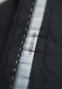 Zwart stoffen detail met fijne stiksels en een zilveren label met de tekst "LINDBERGH." Textuur oppervlak met glans en een matte afwerking.