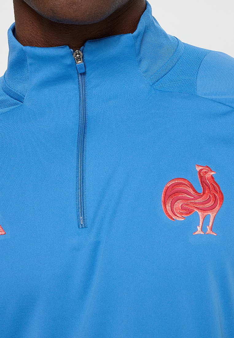Camiseta deportiva azul con cremallera que presenta un alto collar, una pequeña cremallera y un diseño bordado de un gallo rojo en el lado izquierdo del pecho.