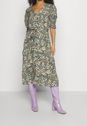 Femme portant une robe midi fleurie avec des manches bouffantes et une ceinture à la taille, associée à des bottes brillantes lavande montant au genou.