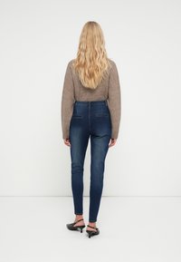 Jeans de tiro alto en azul oscuro con un corte ajustado, que cuentan con bolsillos traseros y detalles sutiles de costura, combinados con un suéter texturizado beige.