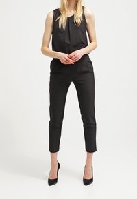 Zwarte mouwloze jumpsuit met een geplisseerde halslijn, slim-fit taps toelopende pijpen en zijzakken; gecombineerd met zwarte hoge hakken.