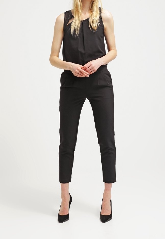 Zwarte mouwloze jumpsuit met een geplisseerde halslijn, slim-fit taps toelopende pijpen en zijzakken; gecombineerd met zwarte hoge hakken.