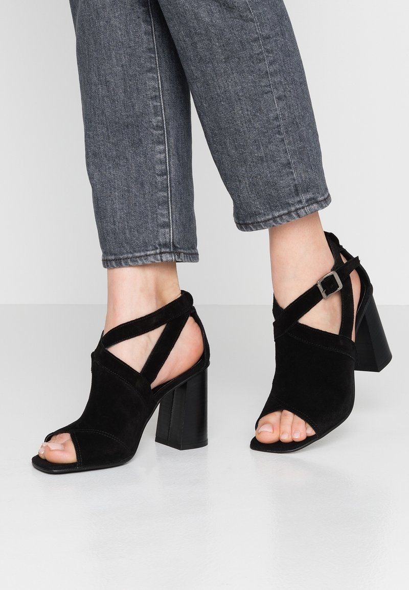 Sandales à talons en daim noir avec bout ouvert, présentant un design à brides croisées et un détail de boucle. Talon épais pour plus de stabilité.