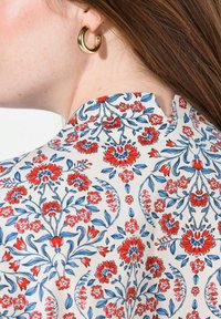 Prenda de diseño floral en crema, con flores rojas y azules. Tejido suave con un pequeño collar muesca. Arete de aro dorado visible.