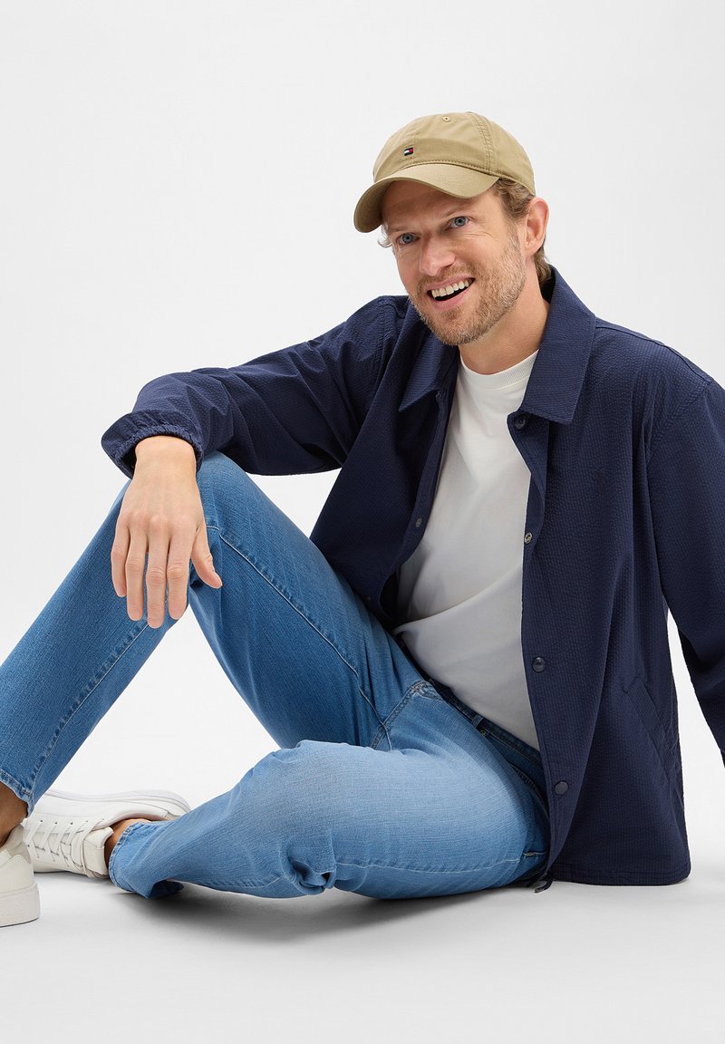 Homme portant une casquette beige, une veste marine, une chemise blanche, un jean bleu clair et des baskets blanches, assis par terre et souriant devant un fond blanc.