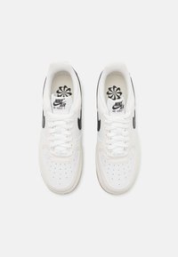 Bílé tenisky Nike Air Force 1 s černými logy swoosh, bílými tkaničkami a perforovanými špičkami, pohled zhora na jednobarevném pozadí.