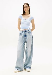 Top azzurro chiaro a coste con maniche corte, colletto ondulato e piccolo dettaglio rosso, abbinato a jeans in denim chiaro a gamba larga e sneakers bianche.