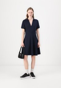 Marineblå kortermet kjole med V-hals, utsvingt design og myk stoff, parret med svarte joggesko og en liten svart veske.