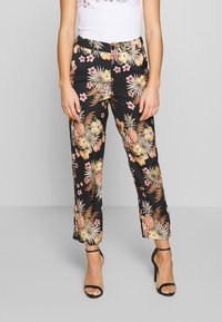 Vrouw in zwarte broek met bloemenprint, witte top en zwarte hoge hakken, staand tegen een effen grijze achtergrond.
