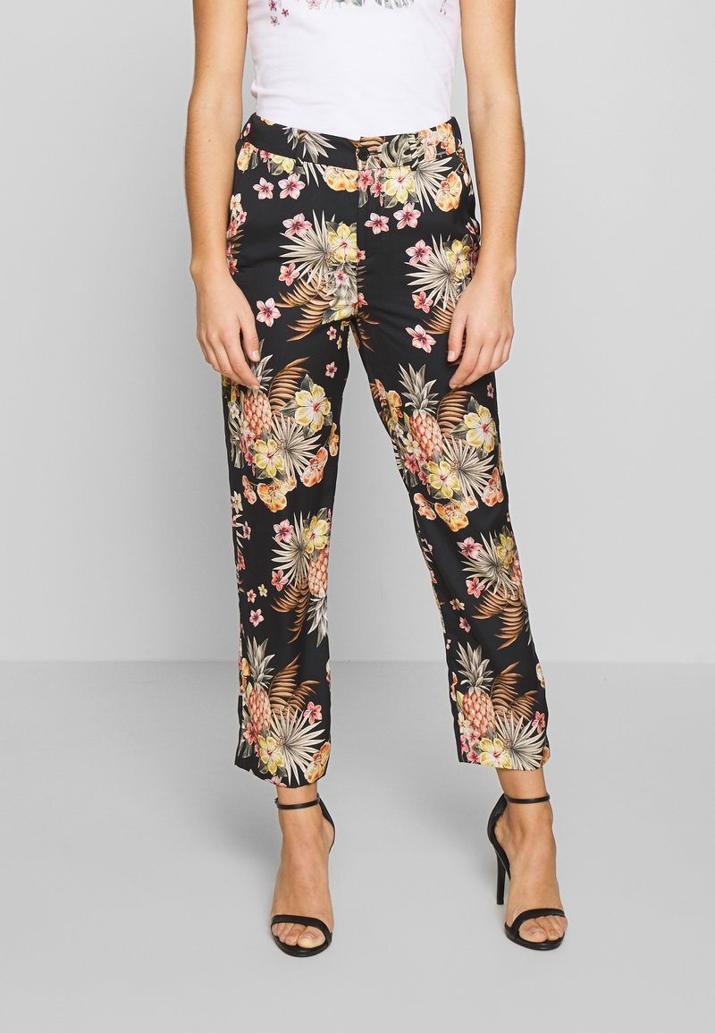Vrouw in zwarte broek met bloemenprint, witte top en zwarte hoge hakken, staand tegen een effen grijze achtergrond.