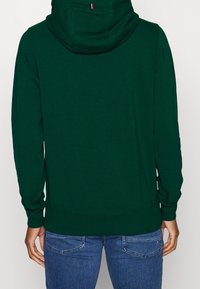 Sudadera verde con un bolsillo tipo canguro, puños y dobladillo acanalados. Presenta una capucha de doble capa con un pequeño acento de marca en la parte trasera.