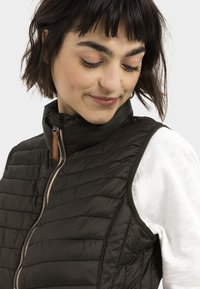 Zwarte, gewatteerde vest met hoge kraag en rits, voorzien van een bruine leren accent. Draagt over een witte lange mouwen shirt met geribbelde manchetten.