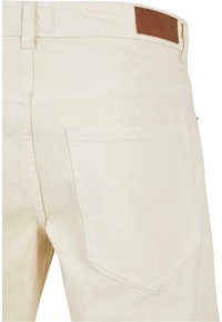 Pantalons en denim beige clair avec une poche arrière et un patch en cuir marron sur la ceinture. Tissu texturé avec un motif de tissage subtil.