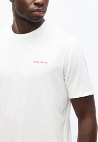 Κοντινό πλάνο ατόμου που φοράει λευκό T-shirt Helly Hansen, δείχνοντας το πάνω μέρος του κορμού και μέρος του προσώπου που κοιτάζει προς τα πλάγια.