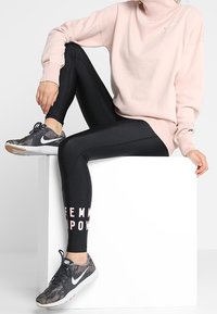 Henkilö istuu valkoisella laatalla, päällään vaaleanpunainen neuletakki ja mustat legginsit, joissa on "FEMME POWER" -teksti, yhdistettynä harmaisiin urheilukenkiin.