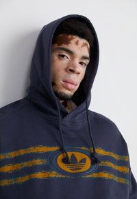 Tengerészkék kapucnis pulóver, húzózsinórral, elhalványult narancssárga és türkiz csíkozott grafikával és középen elhelyezett logóval az elején. Puha szövettextúra.